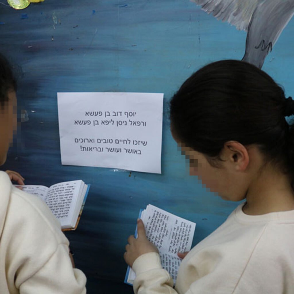 Keren Hayeled students praying near a printed sign with names of orphanage donors to pray for, as part of the orphanage Prayer Project